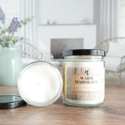 warm mahogany candle is shown with one open laying flat with the top showing, the lid is off to the side. the other candle is standing next to that one with the lid on and showing the label that has trees in the background. there is an image of a fireplace in the background surrounded by 2 wicker chairs. there is also a sage green vase with lavender buds in the back left of the image.