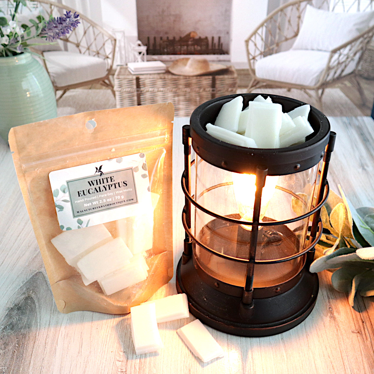 A white eucalyptus wax melt bag is standing next to a lit bronze wax melt burner. this has the white glittery bits of wax in it and there are some more of these scattered along the base. in the background is a fireplace flanked by 2 wicker chairs.