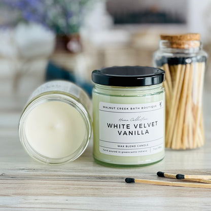 White velvet vanilla candle with lid to the right of another candle with the top off to show the creamy wax inside. there are a few matches scattered in front and a pretty glass jar of matches in the background