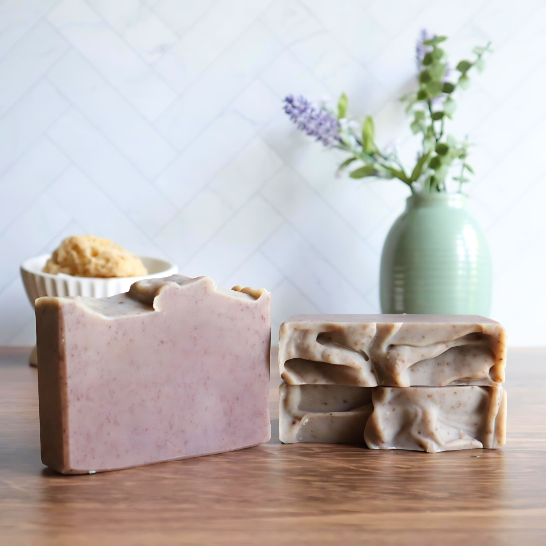 3 wild lavender soaps are shown, one is standing tall to show the purple soaps with speckled purple soap throughout. next to this are 2 more soaps laying flat with the sculpted tops showing. in the background is a sage green vase with flower buds and a marble herringbone backdrop.