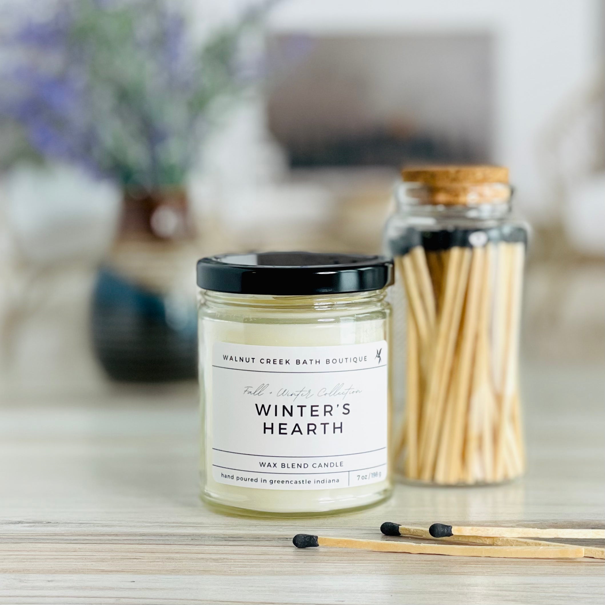 winters hearth candle with black lid. there are a few matches scattered in front and a pretty glass jar of matches in the background