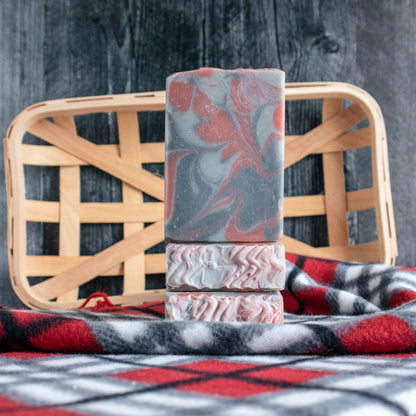2 bars of Cozy Flannel soap laying on a flannel fabric tops facing forward. Another bar is sitting on top of these facing forward showing pretty black, red and grey swirls. There is a blackish background with a wooden tray for interest.