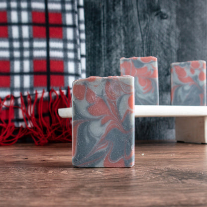 there is a bar of Cozy Flannel soap in for foreground and then 2 more soaps sitting high on a wooden tray in the background The background is black wood with a grey, red and black scarf