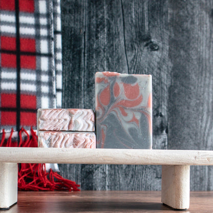 there are 2 bars of Cozy Flannel soaps stacked tops facing forward on a raised wooden trey. There is a 3rd bar standing next to these facing forward. The background is black wood with a grey, red and black scarf
