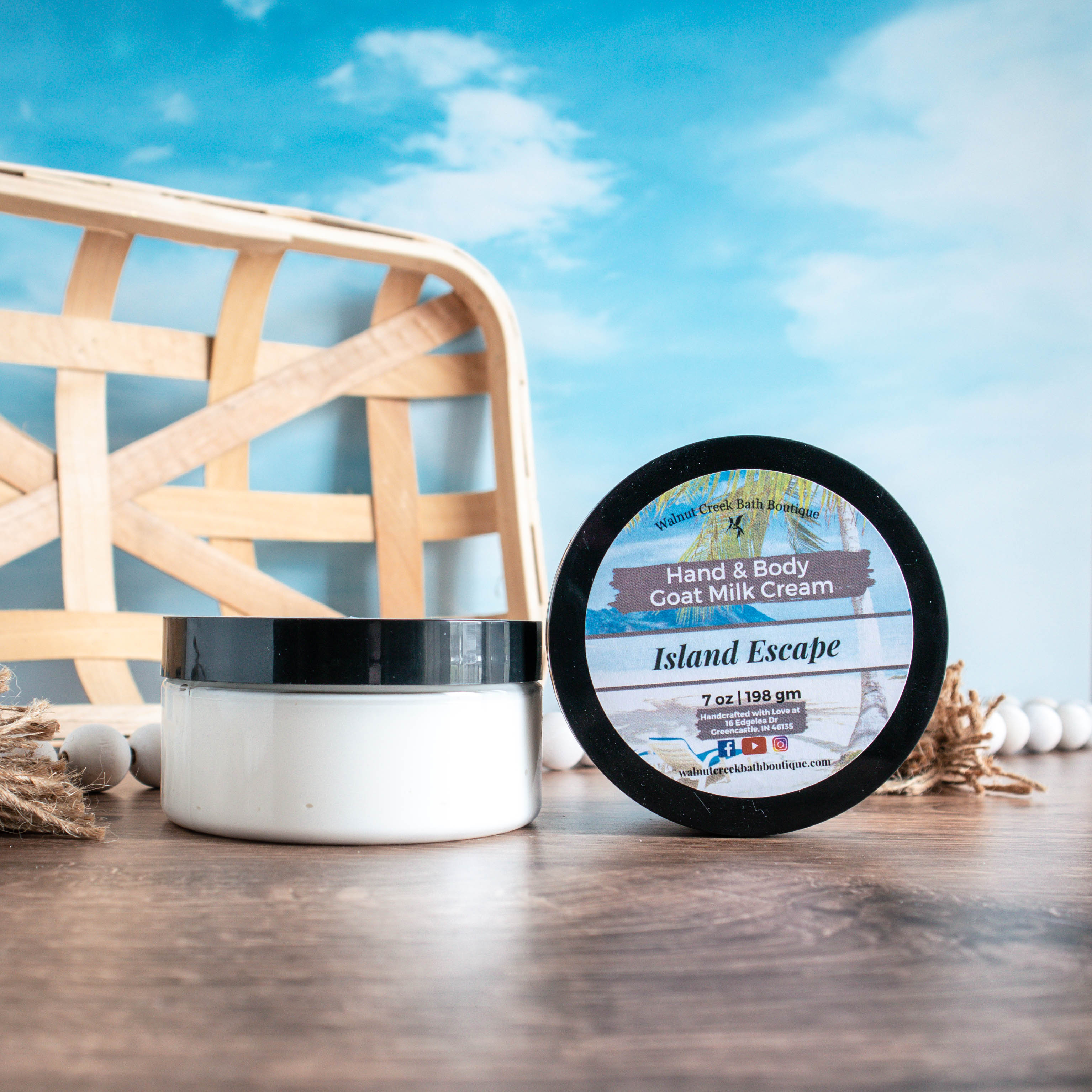 a 7oz  jar of island escape cream is sitting to the left of another jar that is on its side so the top is facing outward. There are white wooden beads in the background along with a wood tray. they are sitting on a warm brown board and there is a sky blue background