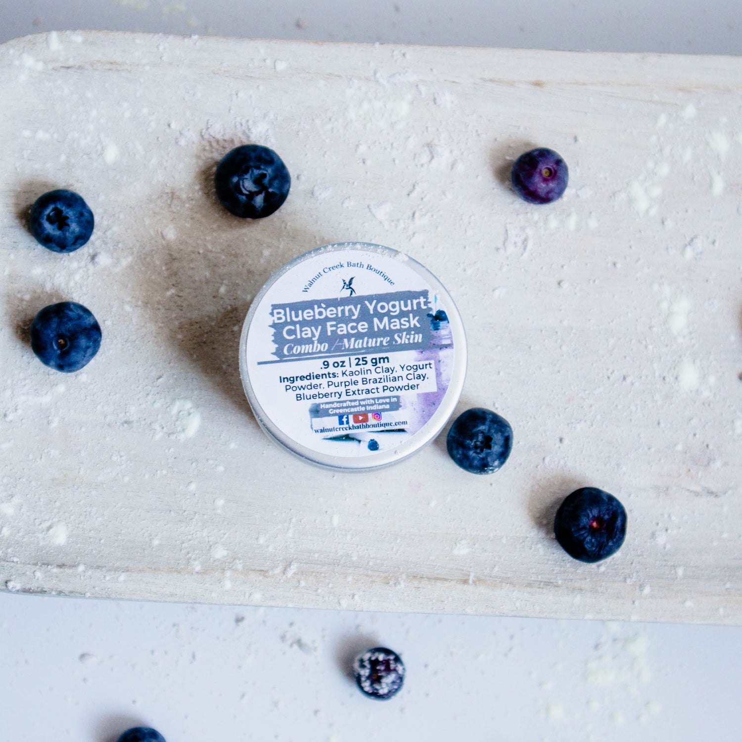 Blueberry yogurt clay face mask is a small glass jar. This is sitting on a wooden board with yogurt powder, clay mask powder and blueberries surrounding it.