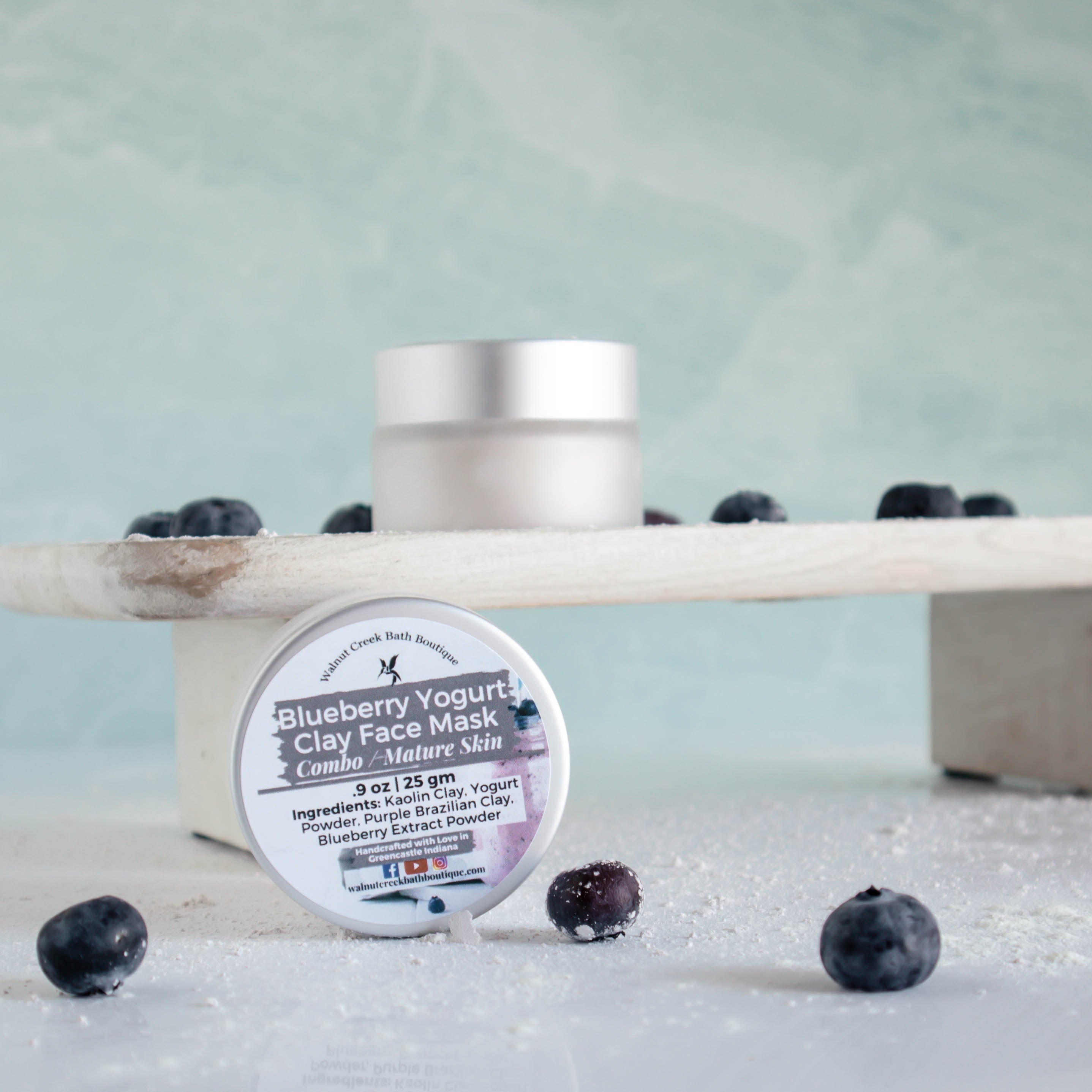 Blueberry Yogurt clay mask jar sitting on its side with another on a wooden board.. There is a dusting of clay mask powder and random blueberries surrounding all the items.