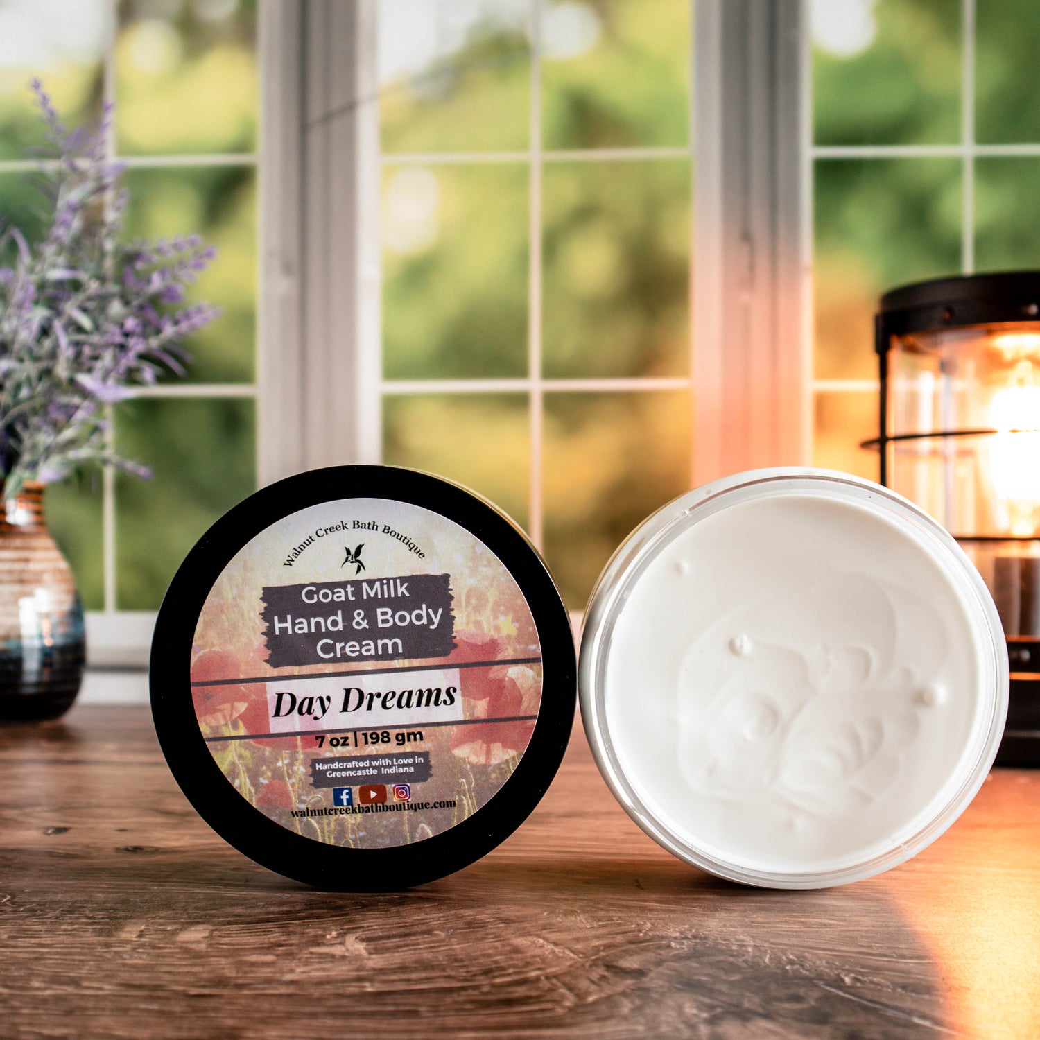 7 oz day dreams cream, one with top showing a pretty label of pink flowers, next to this is an open jar showing contents. There is a lush green background behind a window, a light in the back right and a pretty vase with flowers in the back left.