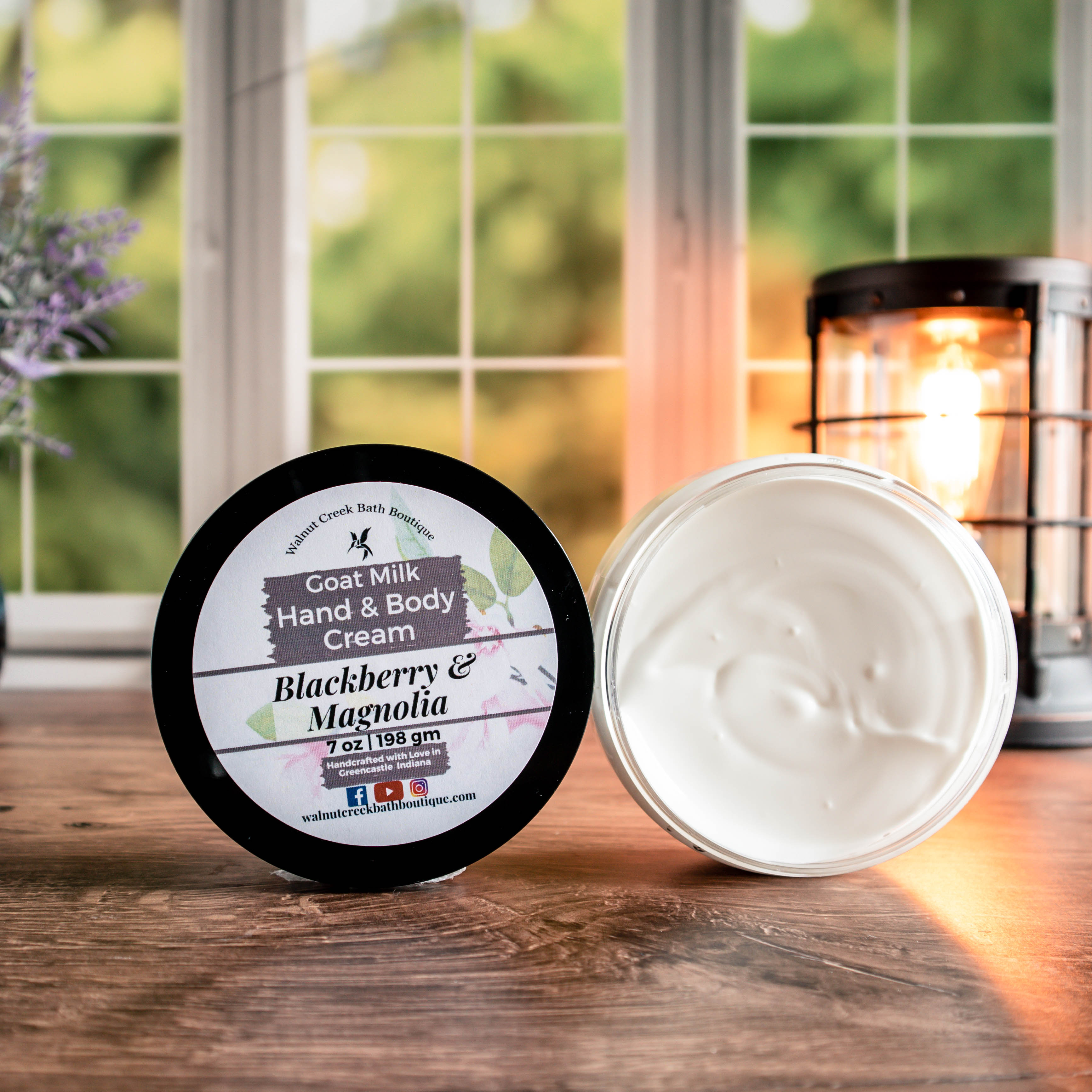 7 oz Blackberry &amp; Magnolia cream, one with top showing a pretty label of pink orchid flowers, next to this is an open jar showing contents. There is a lush green background behind a window, a light in the back right and a pretty vase with flowers in the back left.