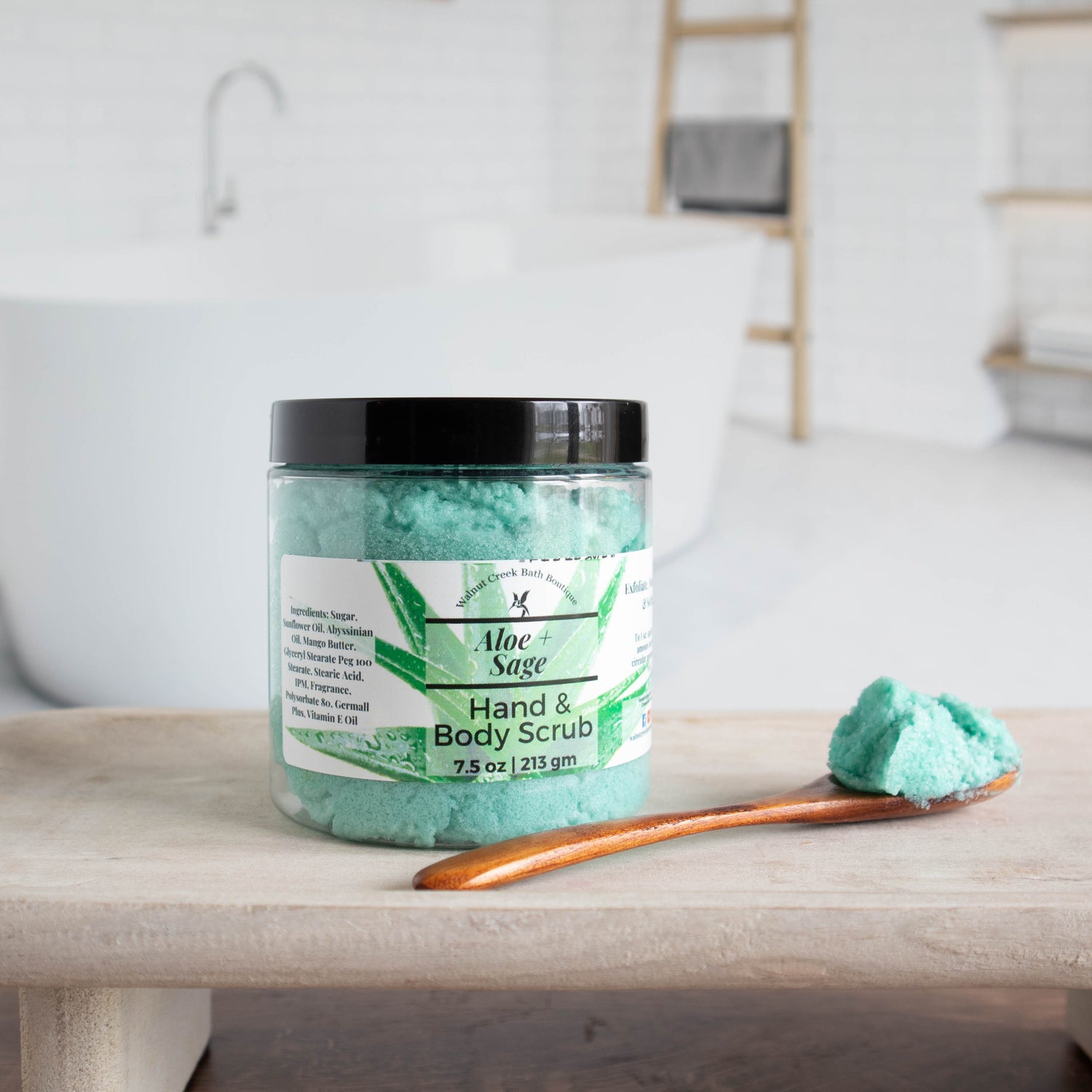 Aloe &amp; Sage scrub sitting on a board, with a wooden spoon with a bit of scrub on it. There is an image of a beautiful bathroom in the background.