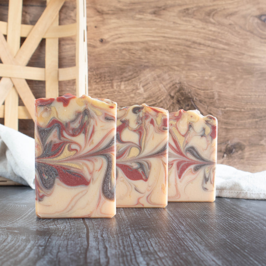 3 susnset soaps lined up in a diagonal. They have a cream base with a rust red, brown and yellow swirl. They are sitting on a black wood base with a walnut wood background. There is a creamy towel in the background as well.