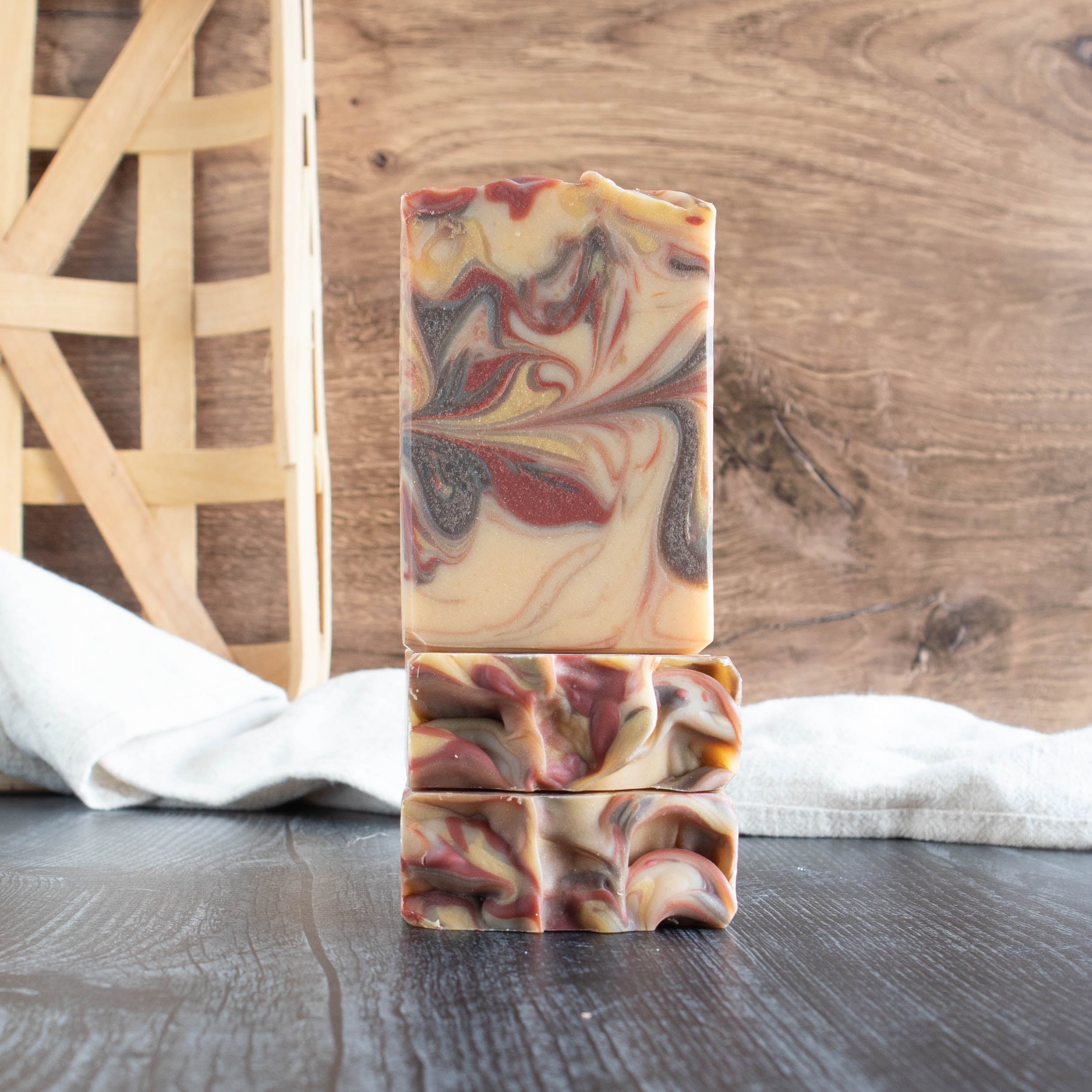 3 susnset soaps. Two are laying flat with the tops facing out and one is sitting on top of them showing the face of the soap. They have a cream base with a rust red, brown and yellow swirl. They are sitting on a black wood base with a walnut wood background. There is a creamy towel in the background as well.