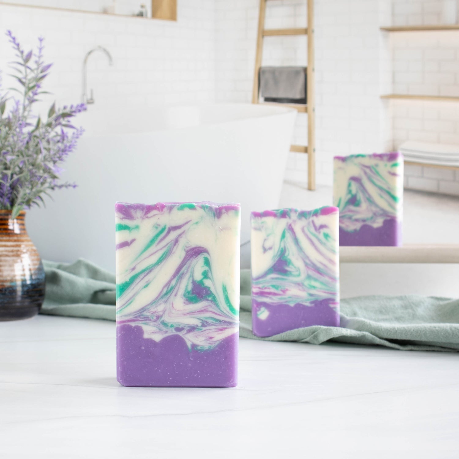 three lavender soaps in a diagonal. They have a purple bottom with a swirled top. The swirls have a white base with 2 purples and a green swirls. They are sitting on a white board with a sage green towel running between them. There is an image of a spa type bathroom in the background