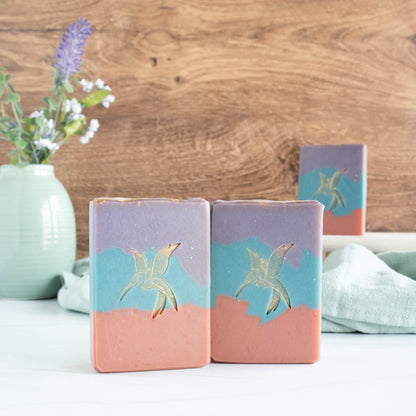 3 sea salt and orchid soaps, 2 towards the front and one in the background. The soaps are rust red on bottom, a blue middle and a purple to. There is also a hummingbird stamp in the middle that is gold. There is a wooden background, a pretty sage green vase in the back left and then a sage green towel running in the background.
