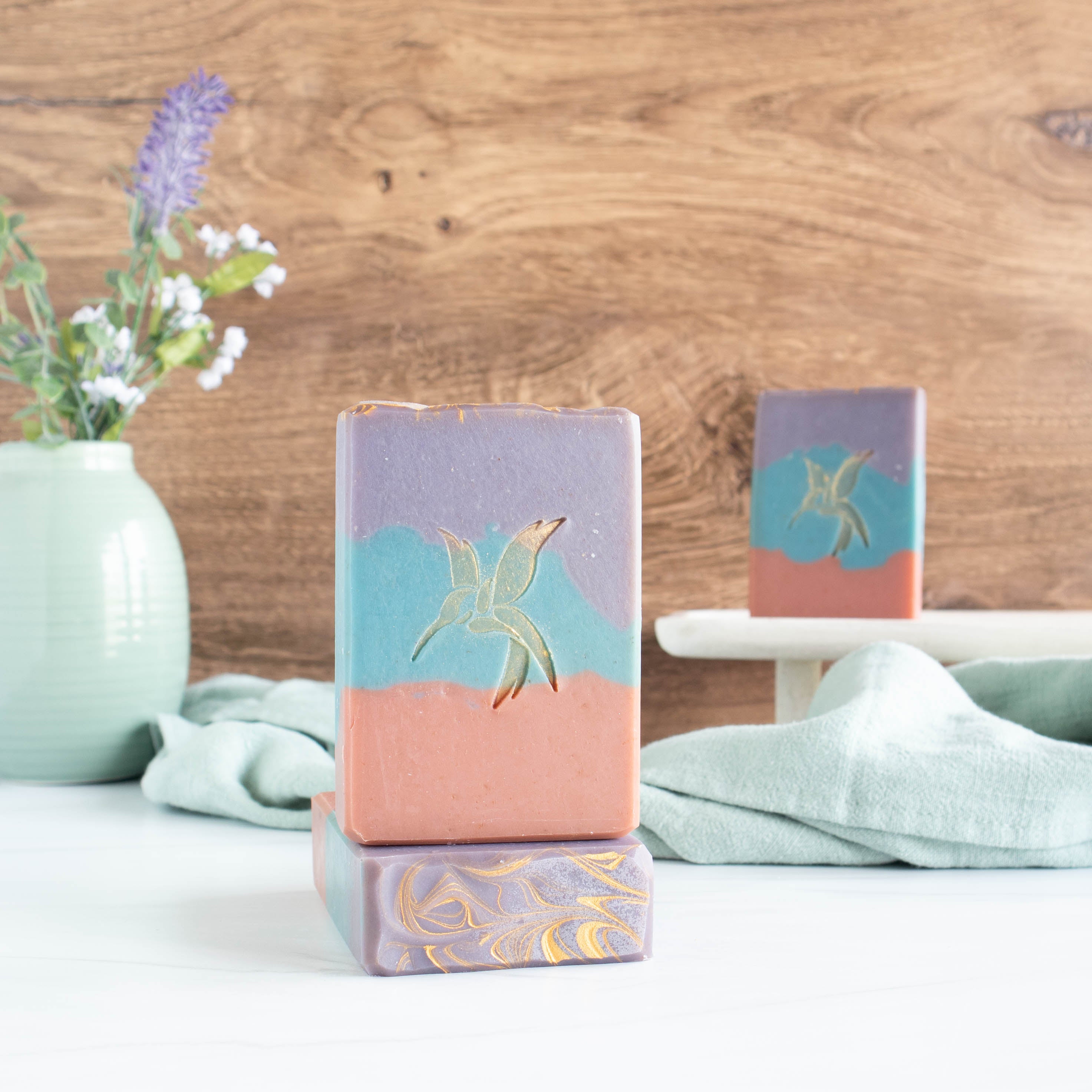 3 sea salt and orchid soaps, 2 towards the front and one in the background. The soaps are rust red on bottom, a blue middle and a purple to. There is also a hummingbird stamp in the middle that is gold. There is a wooden background, a pretty sage green vase in the back left and then a sage green towel running in the background.