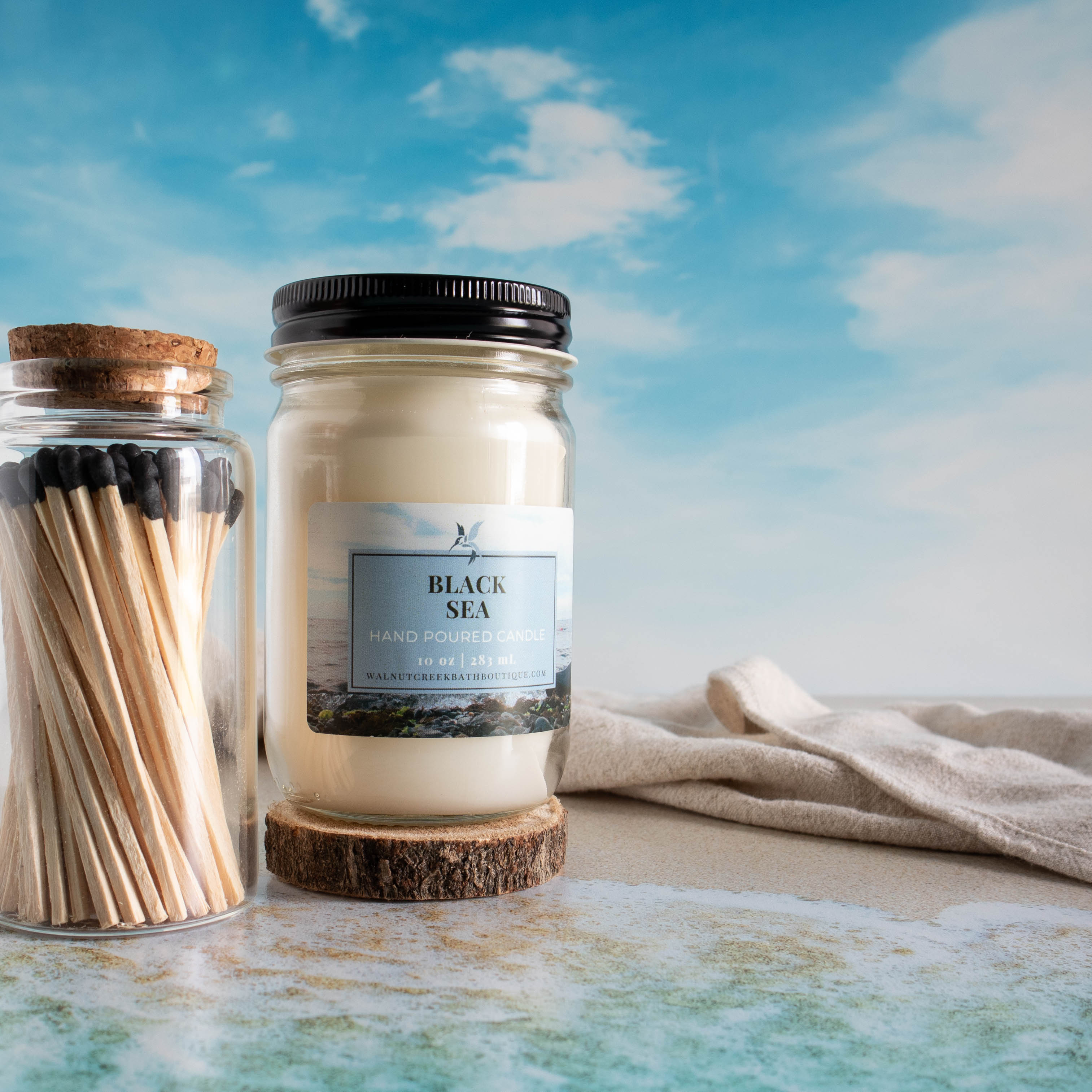 black sea candle with a jar of wooden matches. There is a sky background and a shore line in front of candle