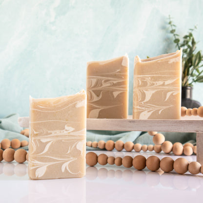 3 aloe and sage soaps with a tan color and a very pretty creamy swirl running through them. One is in the foreground while the other 2 are back a bit standing on a wooden tray. There is a lovely sage green background along with a wooden bead that is running along between the soaps.