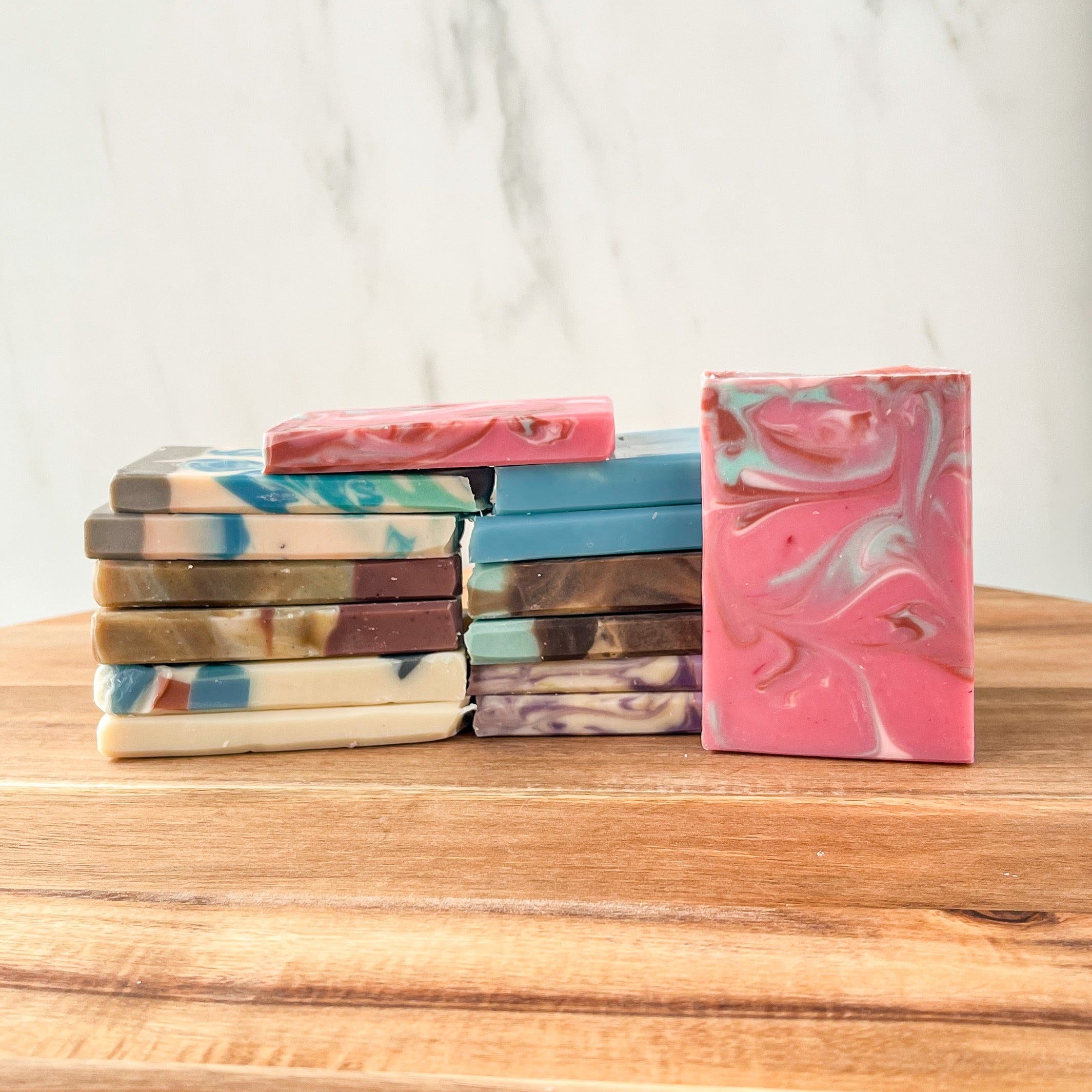 there are 14 different soap samples, 6 are stacked one one side and six on the other. they are facing each other and touching. There is another soap that is on top of these bridging them and the last soap standing off to the right leaning on the right stack.. They are sitting on a wooden board and there is a white background.