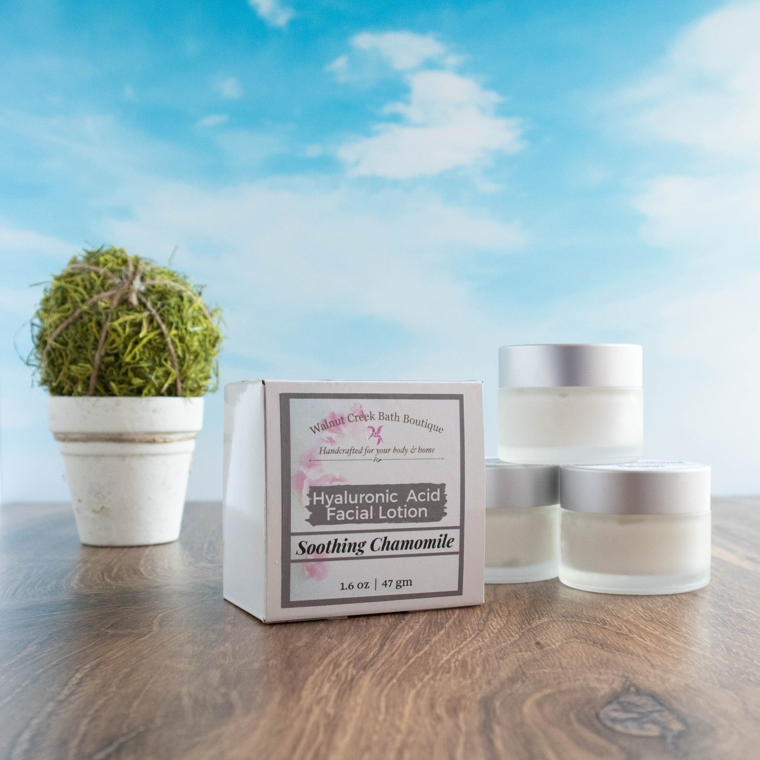 there are 3 hyaluronic acid face cream jars stacked to the right and slightly in front of this is a box which is the outer package of the product. there is a stone vase with a greenery ball. everything is sitting on a wood board and there is a sky background.