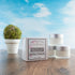 there are 3 hyaluronic acid face cream jars stacked to the right and slightly in front of this is a box which is the outer package of the product. there is a stone vase with a greenery ball. everything is sitting on a wood board and there is a sky background.