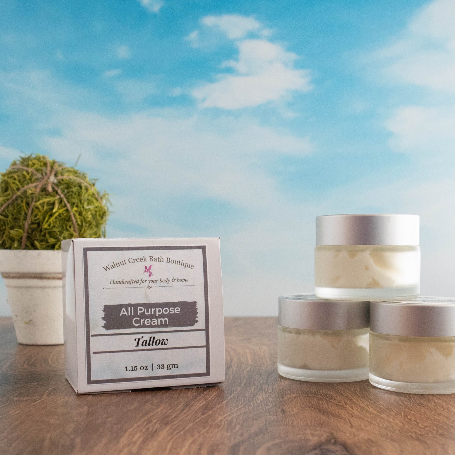 there are 3 tallow face cream jars stacked to the right and slightly in front of this is a box which is the outer package of the product. there is a stone vase with a greenery ball. everything is sitting on a wood board and there is a sky background.