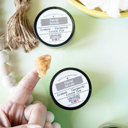 there are 2 jars of face scrub on a green surface with a wooden bead rope. The main focus is on the hand in the bottom left that has a scoop of the scrub on the finger showing the lovely texture of the scrub.