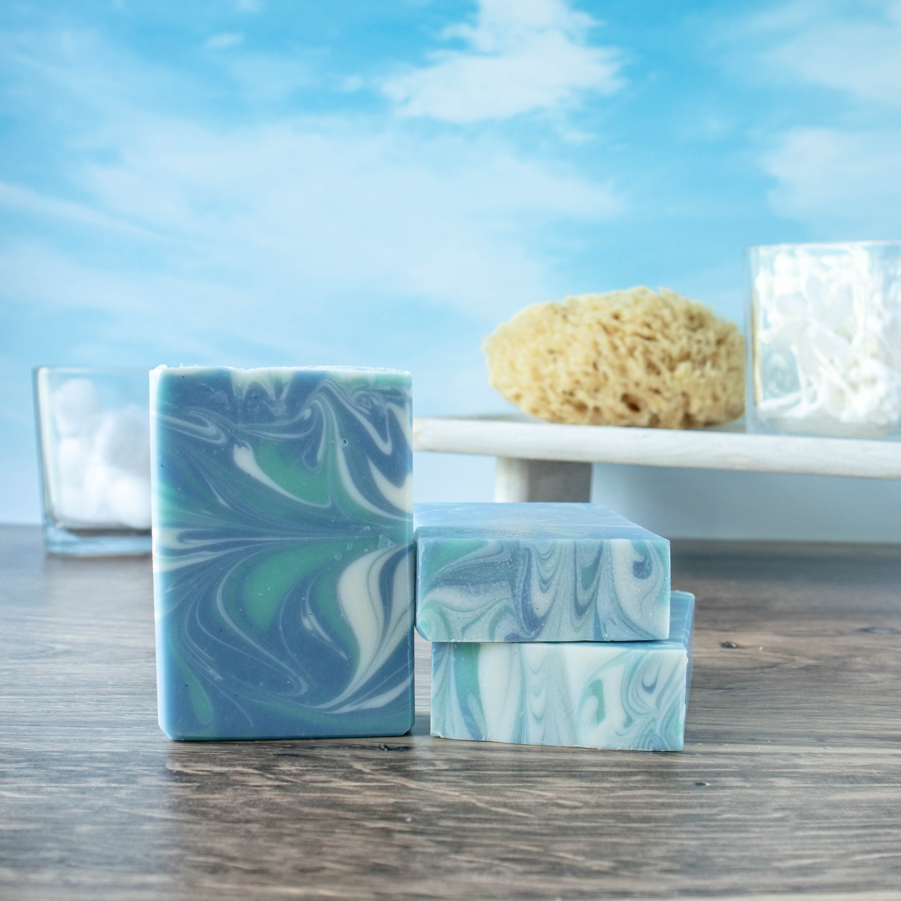 the face on view of tranquil waters, a mostly blue soap with whispy green and white swirls. There are 2 other bars showing a pretty swirl top. There is a sky background with a sea loofah and q-tips on a wooden tray.