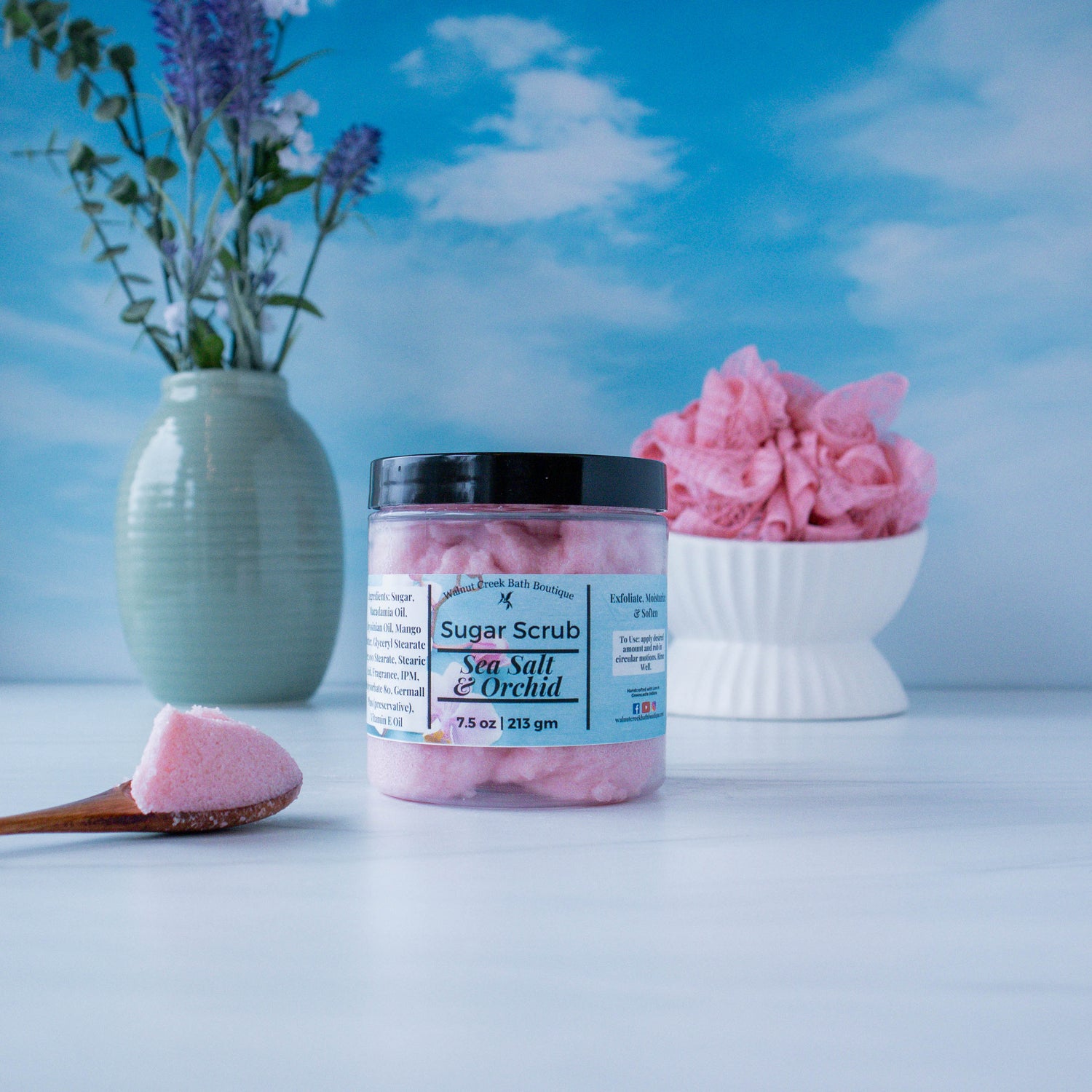 Sea Salt &amp; Orchid scrub is sitting center with a wooden spoon holding some product to show texture.  to the left is a green vase with some greenery and to the right back is a white dish holding a pink loofa. this is all sitting on a white board and there is a sky backdrop.