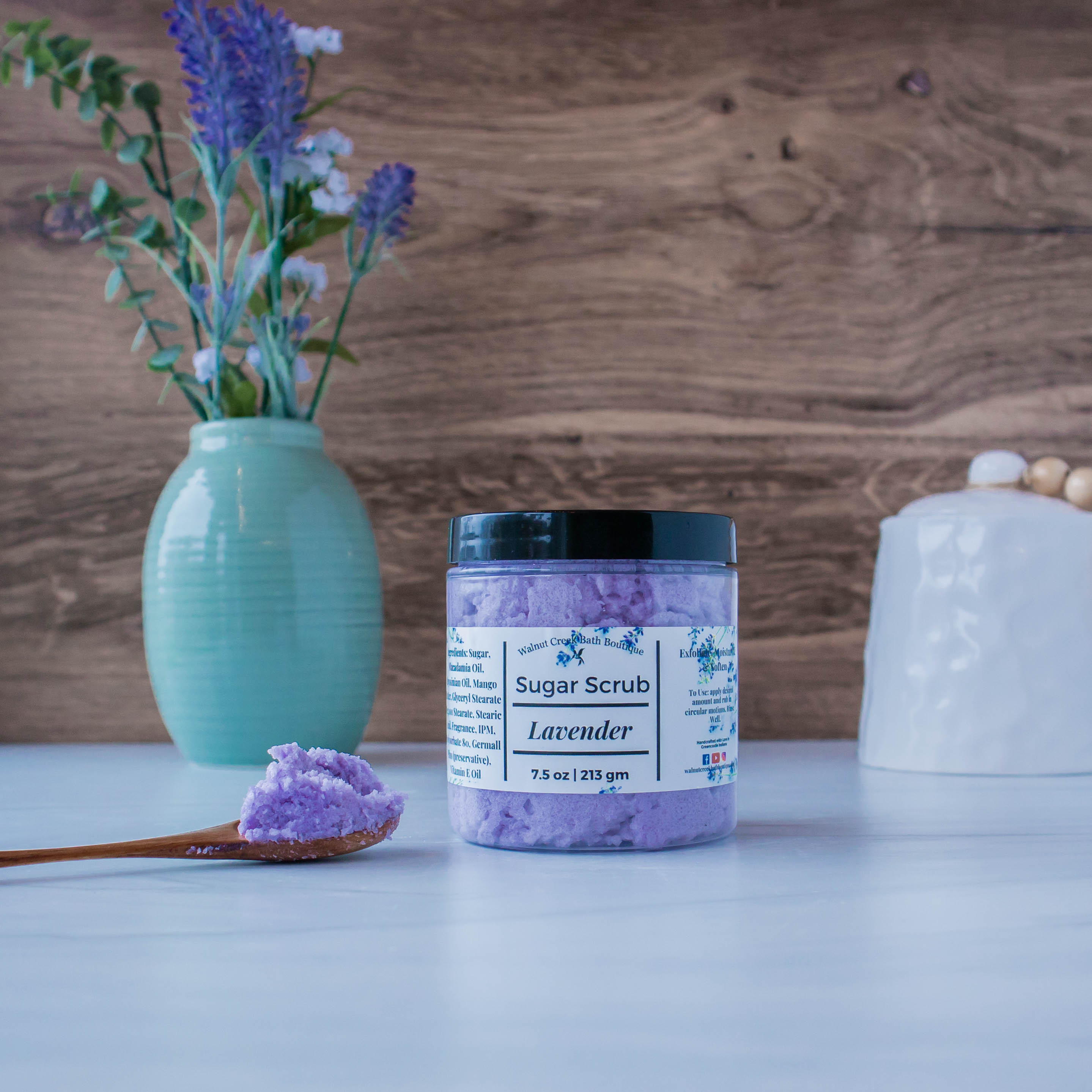 Lavender scrub is sitting center. there is a small wooden spoon holding some of the scrub to show texture. to the left is a blue vase with some greenery and to the right back is a wooden pumpkin. this is all sitting on a white board and there is a brown wood backdrop.