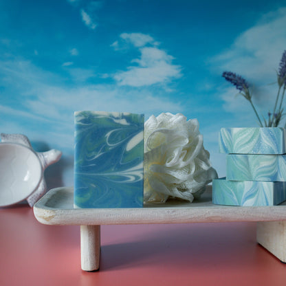 the face on view of tranquil waters, a mostly  blue soap with whispy green and white swirls. There are three other bars showing a pretty swirl top. They are sitting on a raised wood tray with a white loofah. There is a sky background.