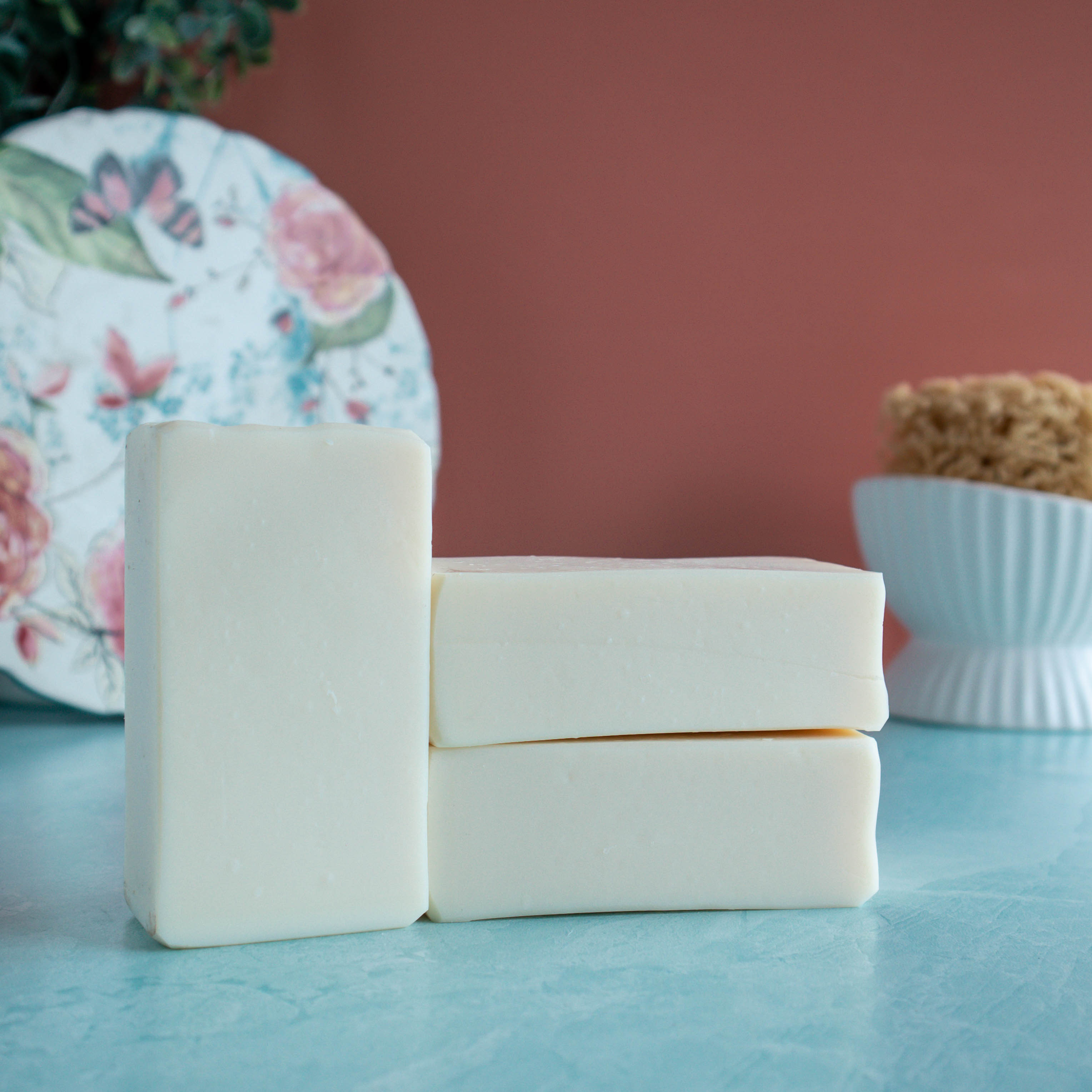 3 bars of creamy white soap are in the foreground, 2 stacked on top of each other. there is a pretty floral plate in the back left corner and a sponge in a white dish in the back right. The background is a lovely coral color and a seafoam green base. 