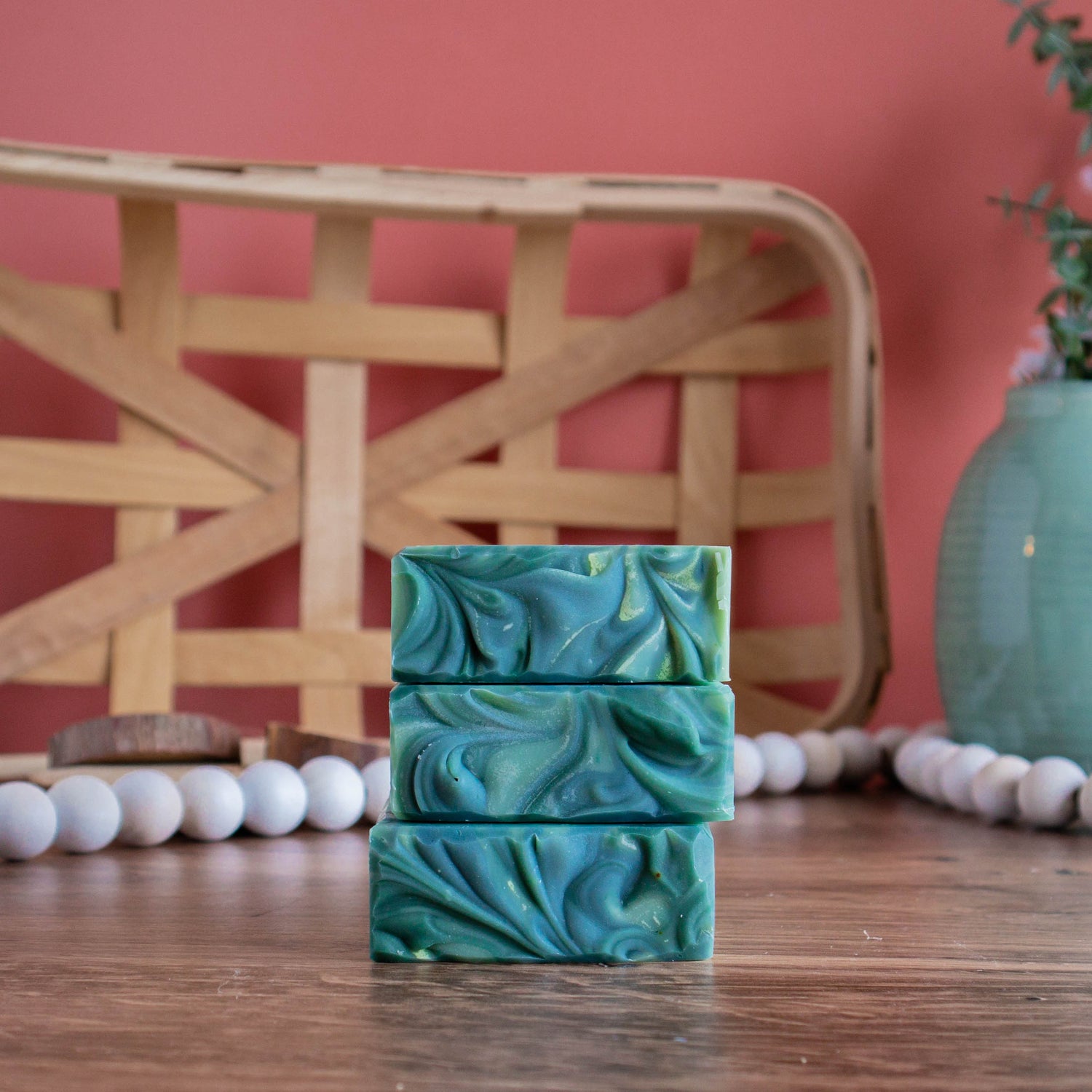 three bars of aspen soap are stacked showing the tops. they are sitting on a wooden board and there is a coral color background. in the back left is a woven wooden basket and in the back right is a green vase with some florals. there is a white bead rope running along in behind the soaps