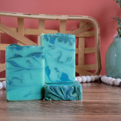 three bars of aspen soap, one laying flat with the top facing with another standing on top of it.  The third is standing next to these. they are sitting on a wooden board and there is a coral color background. in the back left is a woven wooden basket and in the back right is a green vase with some florals. there is a white bead rope running along in behind the soaps