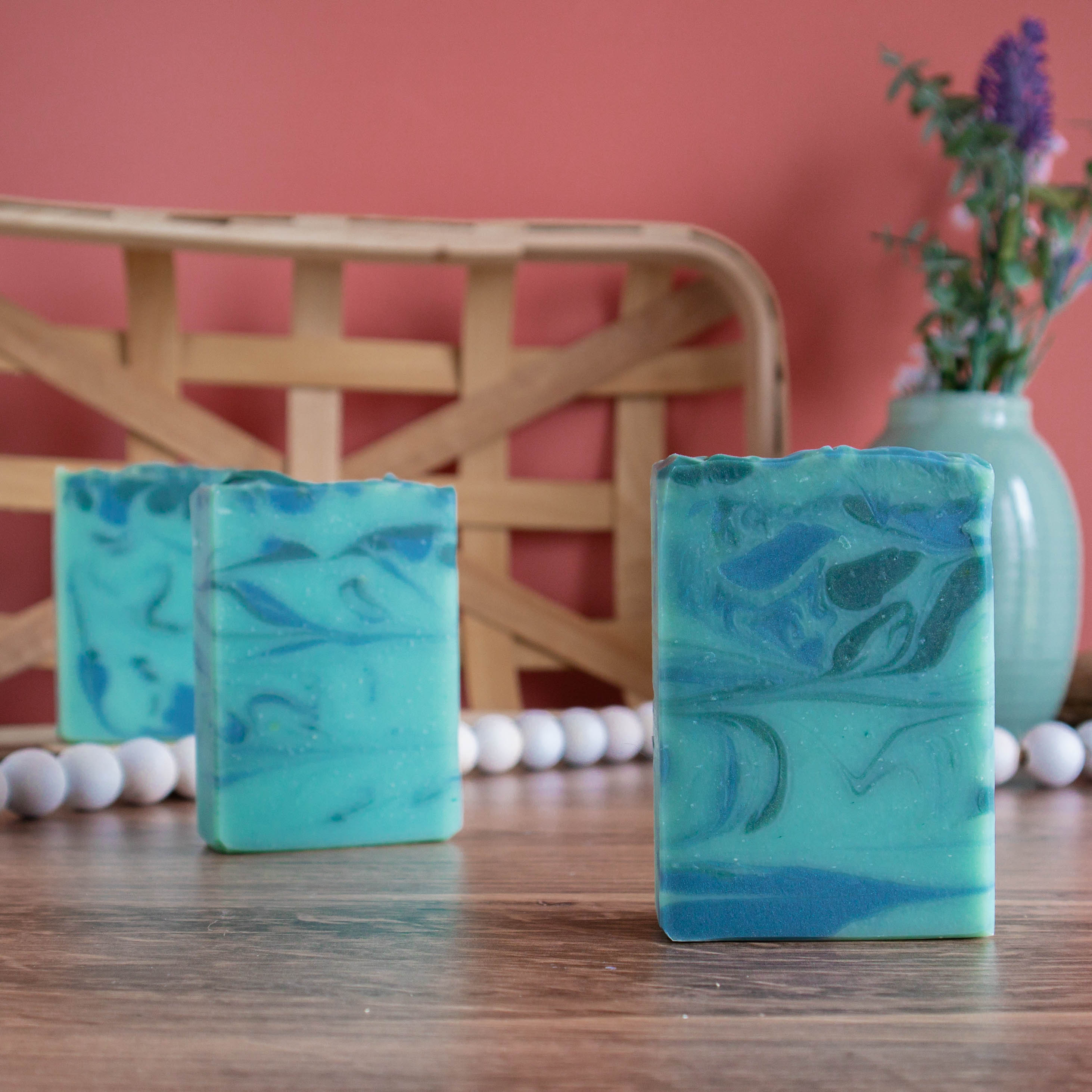 three bars of aspen soap are lined up diagonally left to right. they are sitting on a wooden board and there is a coral color background. in the back left is a woven wooden basket and in the back right is a green vase with some florals. there is a white bead rope running along in between the soaps.