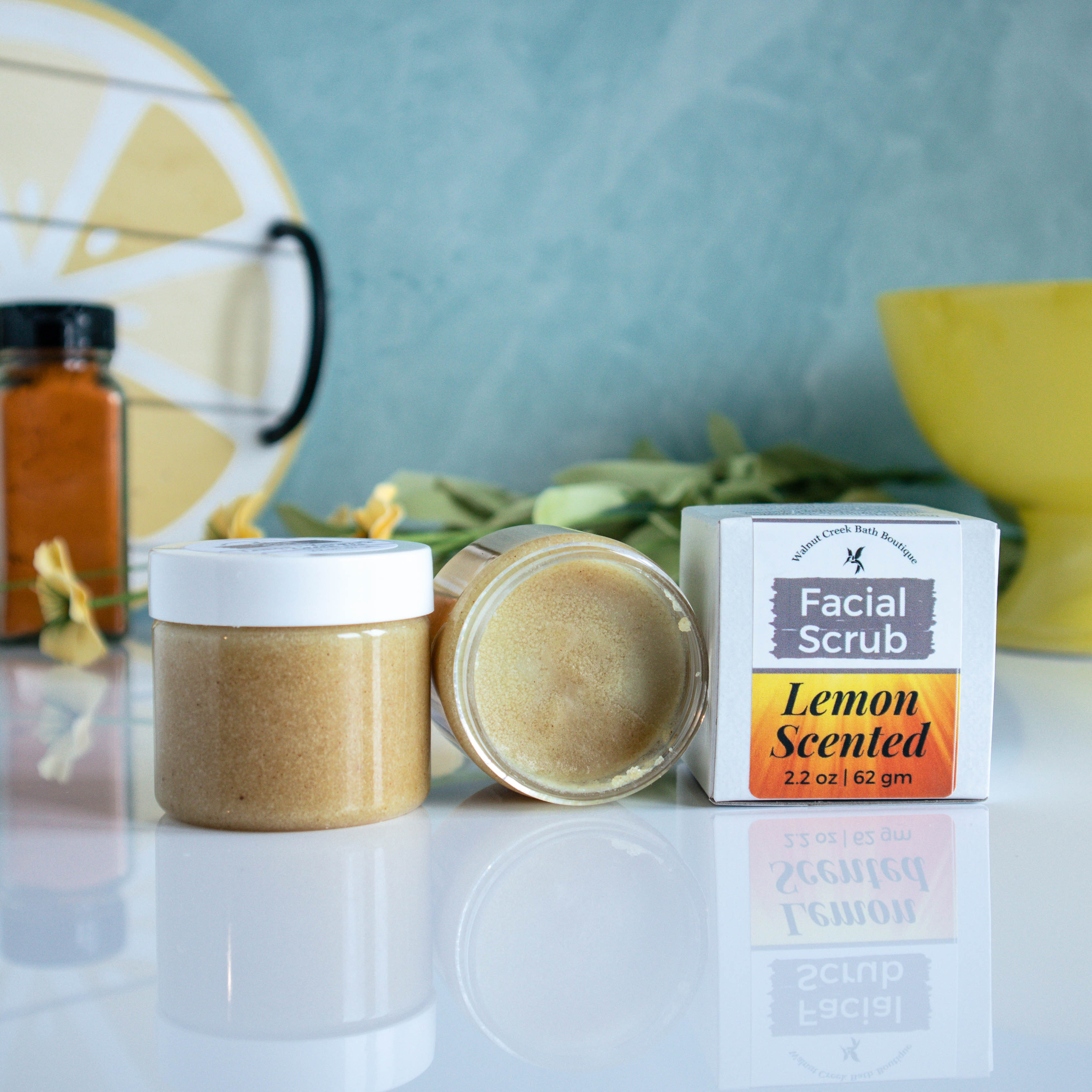 there is one jar with a white lid , next to that is an open jar on its side showing contents of face scrub. Next to that is the outer box with the label of facial scrub, lemon scented facing you. This is sitting on a shinny white  base. There is a blue/green background with a lemon tray off to the back left and a yellow bowl to the back right. There is a jar of turmeric in the background.