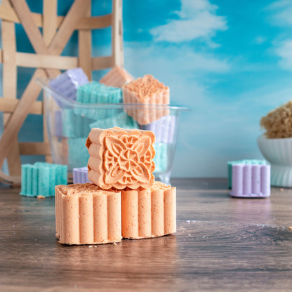 Bright and Sunny shower steamers stacked. One facing showing a pretty scroll on top of the orange steamer. There is a mound of steamers in the background. There is a pretty blue sky in the background.