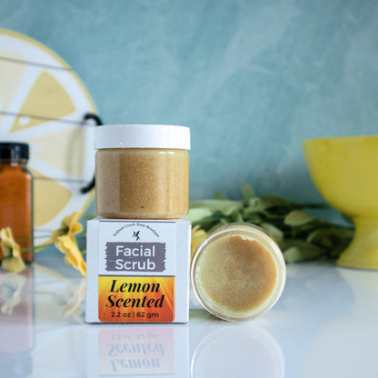 there is one jar with a white lid sitting on the box showing the label with the words facial scrub, lemon scented facing out.  Next to that is an open jar on its side showing contents of face scrub.  This is sitting on a shinny white base. There is a blue/green background with a lemon tray off to the back left and a yellow bowl to the back right. There is a jar of turmeric in the background.