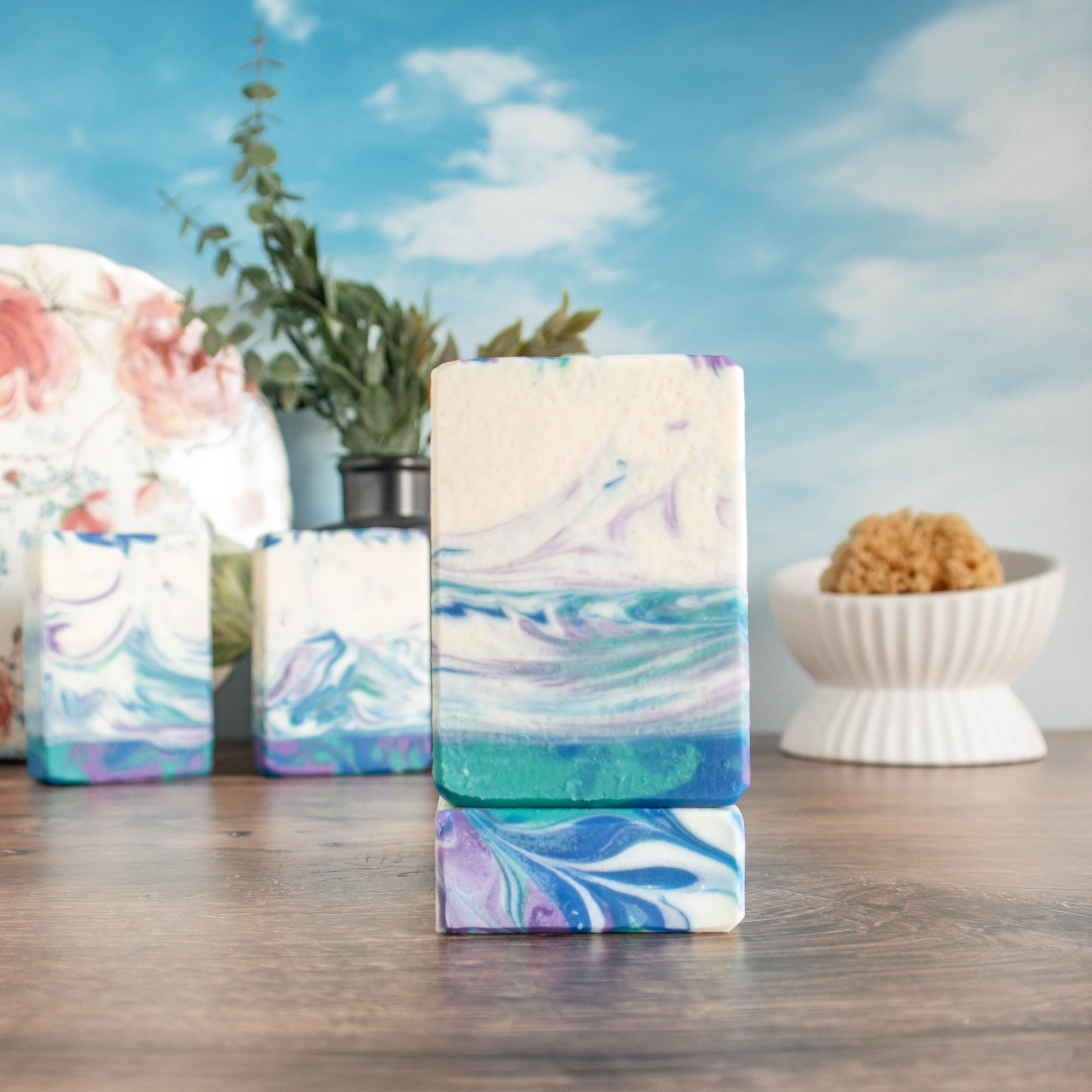 Irish Rains soap, one showing the face and the very pretty colors of purple green and blue swirls standing on another to show the swirled top. Two more are in the back left in front of a floral plate. In the back right is a white soap dish with a loofah There is a pretty sky background.