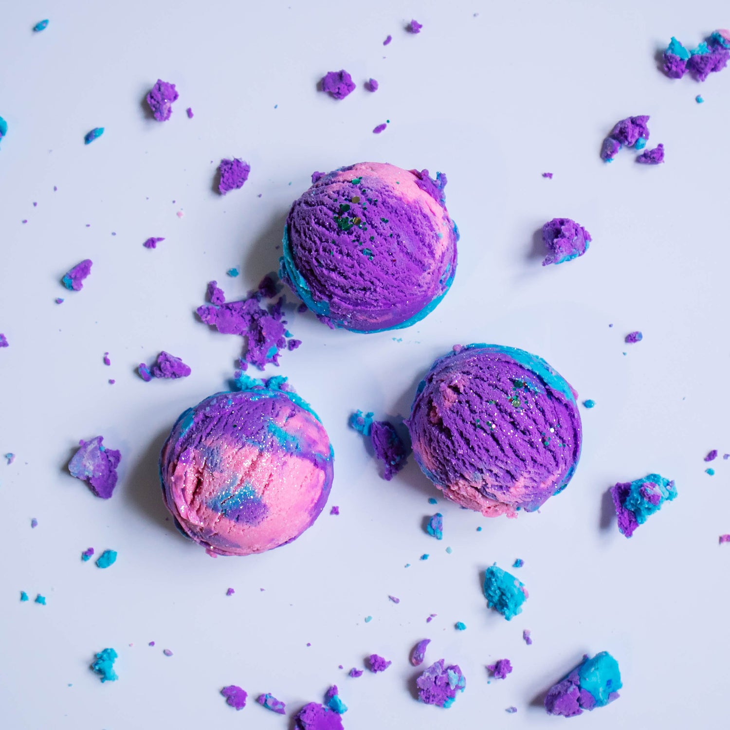 In this top down view there are 3 bubble scoops showing the purple, pink and blue swirls and the glitter that is on top. There are bubble bath crumbles scattered around the white surface.