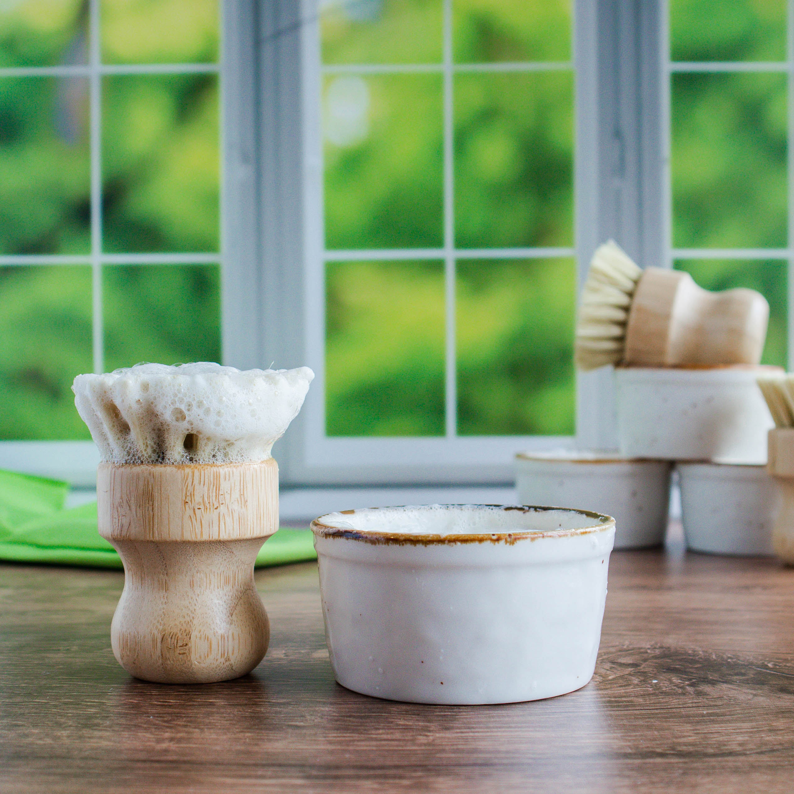 there is a bamboo dish brush full of lather standing next to a crock. in the background on the right is a trio of crocks stacked with one bamboo brush laying across the top and another standing next to them. there is a window in the background showing a blurring of green leaves.