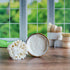 there is a bamboo dish brush full of lather laying next to a crock that is tipped on its side to show the lather in the crock.. in the background on the right is a trio of crocks stacked with one bamboo brush laying across the top and another standing next to them. there is a window in the background showing a blurring of green leaves.