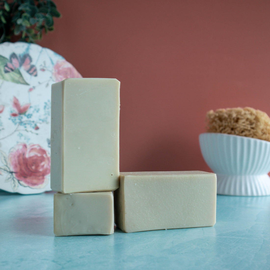 3 bars of creamy tan soap are in the foreground, 2 stacked on top of each other. there is a pretty floral plate in the back left corner and a sponge in a white dish in the back right. The background is a lovely coral color and a seafoam green base.