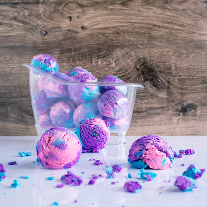 In the background there is a clear toy bath tub filled to the brim with very colorful bubble scoops. In front of this are 3 bubble scoops on their sides facing you showing the purple, pink and blue swirls and the glitter that is on top. There are bubble bath crumbles scattered around the surface.