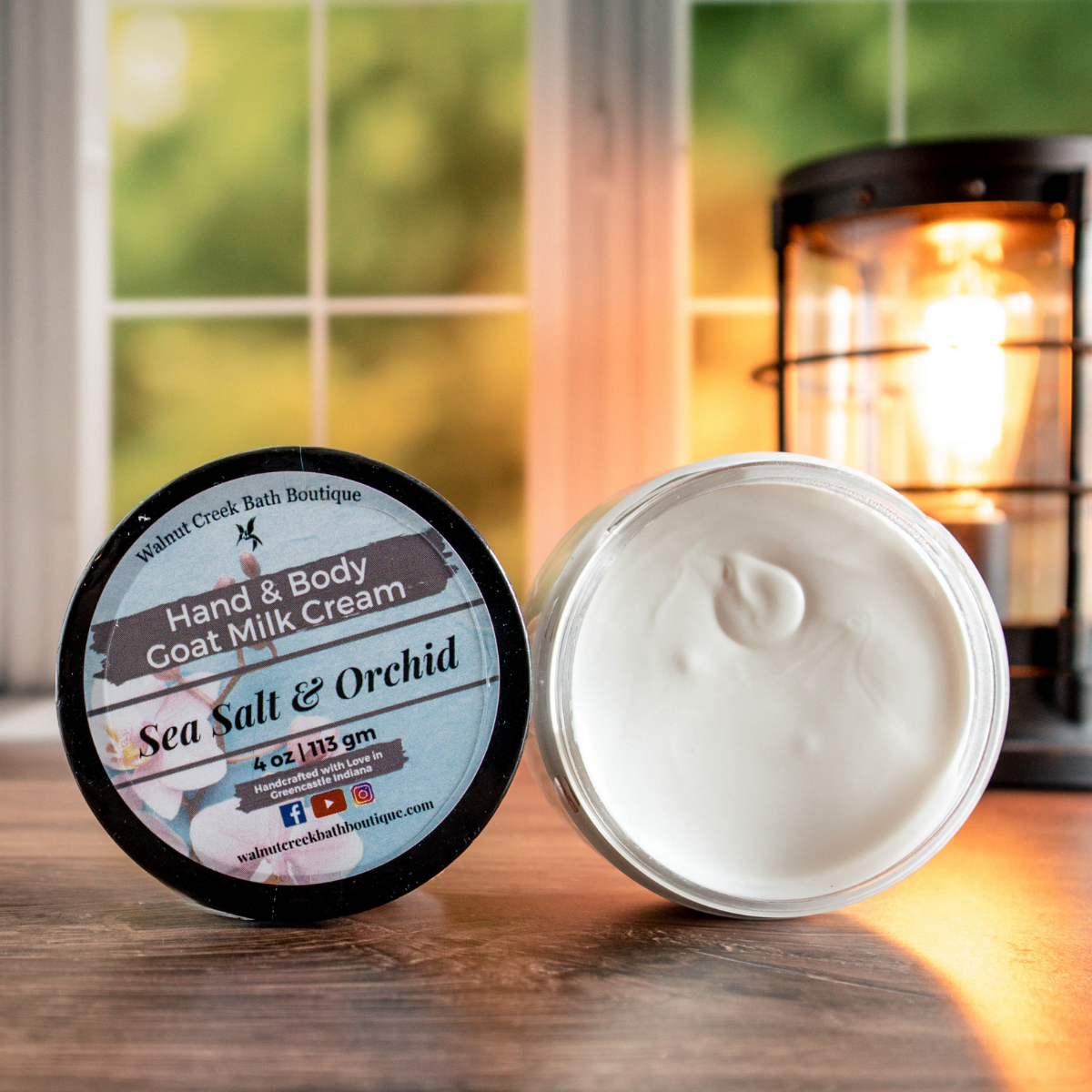 4 oz Sea Salt &amp; Orchid cream, one with top showing a pretty label of pink orchid flowers, next to this is an open jar showing contents. There is a lush green background behind a window and a light in the back right.